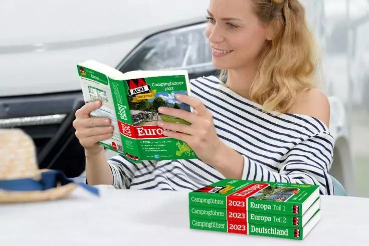 Frau im gestreiften Oberteil liest einen gr&uuml;nen Reisef&uuml;hrer mit dem Titel &bdquo;Europa&ldquo;; zwei gr&uuml;ne Reisef&uuml;hrer liegen gestapelt auf dem Tisch; im Hintergrund ist ein Auto