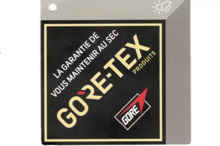 Schwarzes quadratisches Etikett auf grauer Pappe mit goldener Umrandung; diagonal großer goldener Schriftzug GORE-TEX; darüber weiße französische Schrift LA GARANTIE DE VOUS MAINTENIR AU SEC; unten rechts kleines rotes und schwarzes Kästchen mit GORE