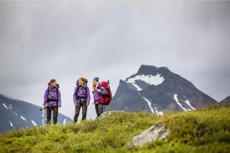 Drei Personen mit Rucksäcken stehen auf einem grasbewachsenen Hang vor Bergen mit Schneeflecken unter bewölktem Himmel