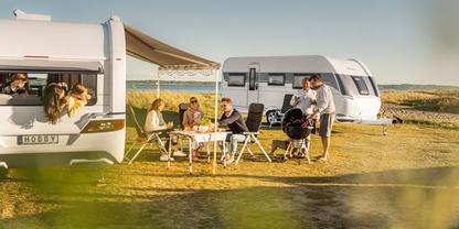Mehrere Personen sitzen an einem Campingtisch unter einer Markise zwischen zwei Wohnwagen; eine Person steht an einem Grill, ein Hund liegt neben dem Tisch; offene Graslandschaft und Himmel im Hintergrund