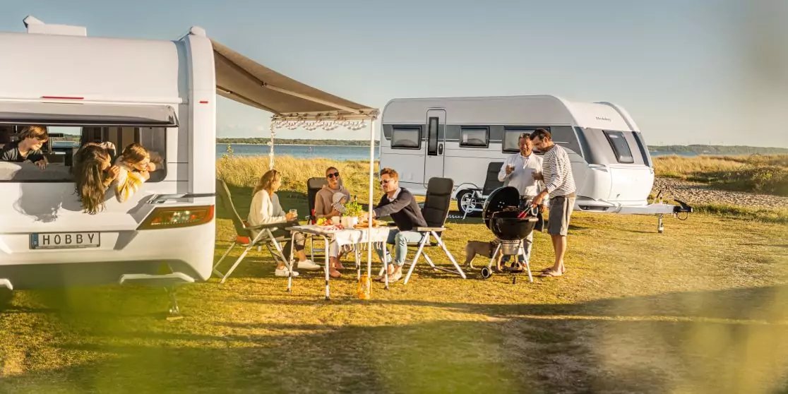 Mehrere Personen sitzen an einem Campingtisch unter einer Markise zwischen zwei Wohnwagen; eine Person steht an einem Grill, ein Hund liegt neben dem Tisch; offene Graslandschaft und Himmel im Hintergrund