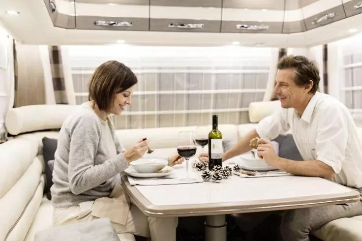 Zwei Personen sitzen an einem Tisch in einem Wohnmobil; auf dem Tisch Teller, Besteck, zwei Weingläser und eine Weinflasche; helle Polster und Vorhänge im Hintergrund