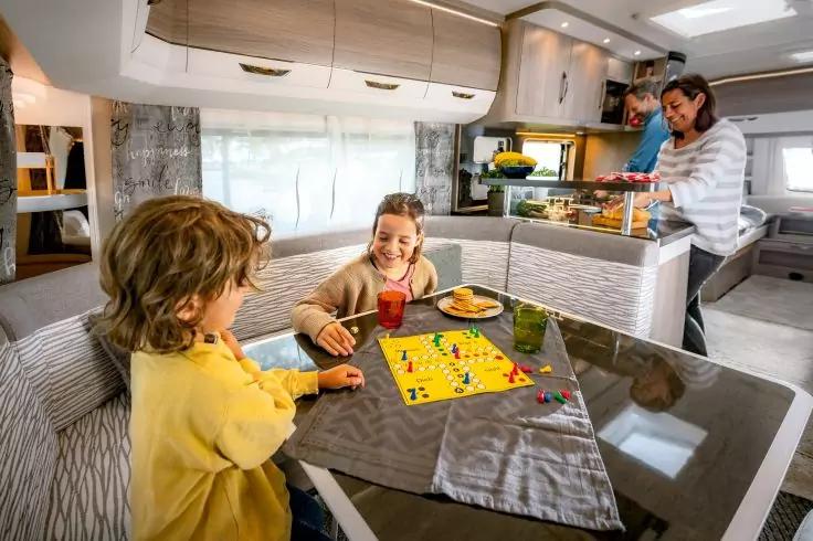 Innenraum eines Wohnmobils: ein Tisch mit Brettspiel, Spielsteinen, Getränken und Gebäck; zwei Personen sitzen am Tisch und bewegen Spielsteine; im Hintergrund zwei Personen an der Küchenzeile, eine hält ein Tablett