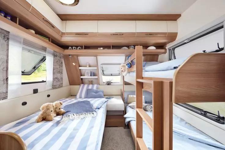 Innenraum eines Wohnmobils mit Etagenbett rechts, Einzelbett links mit blau-weiß gestreifter Bettwäsche und zwei Teddybären, Holzverkleidung und Fenster