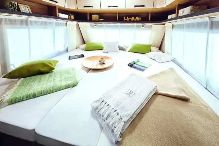 Großes Bett mit weißen Auflagen, beige Decke mit Fransen, grünen Kissen, runder Holzplatte mit Tasse, halbtransparenten Vorhängen an den Seitenfenstern und Regalfächern über dem Bett