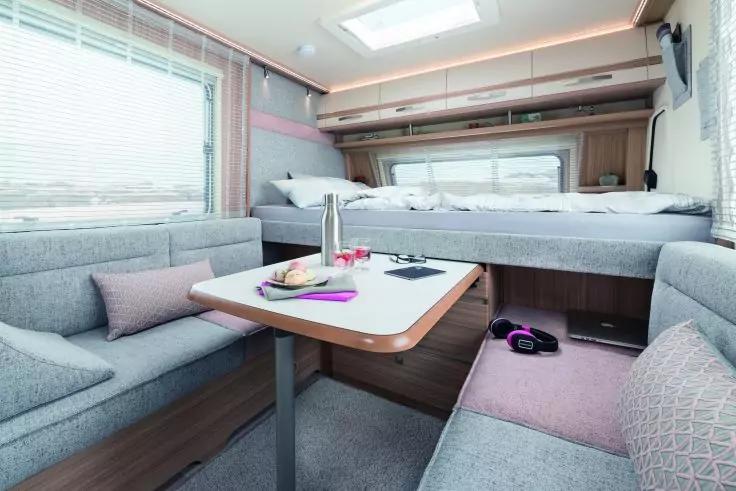 Innenraum mit U-förmiger Sitzbank und Tisch; auf dem Tisch Teller, Tasse und Zeitschrift; Kissen auf den Sitzbänken; erhöhte Liegeflächen über dem Sitzbereich; Fenster mit Jalousien und Dachluke; Kopfhörer auf dem Boden