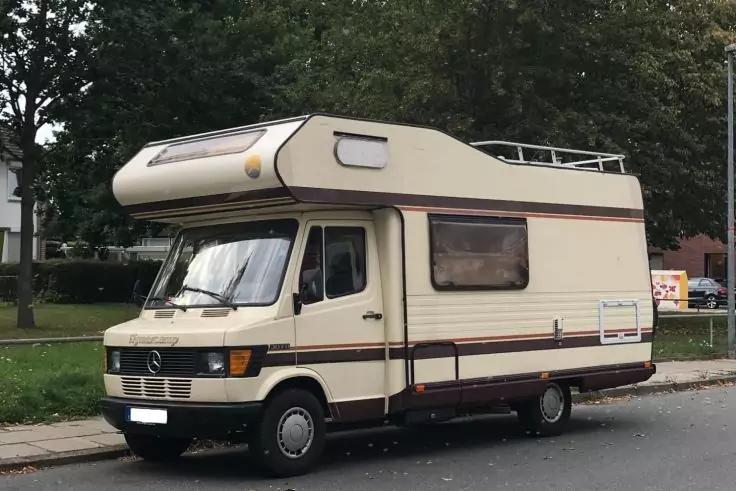 Beiges Wohnmobil mit Alkoven &uuml;ber der Fahrerkabine, geparkt am Stra&szlig;enrand; B&auml;ume und Rasen im Hintergrund