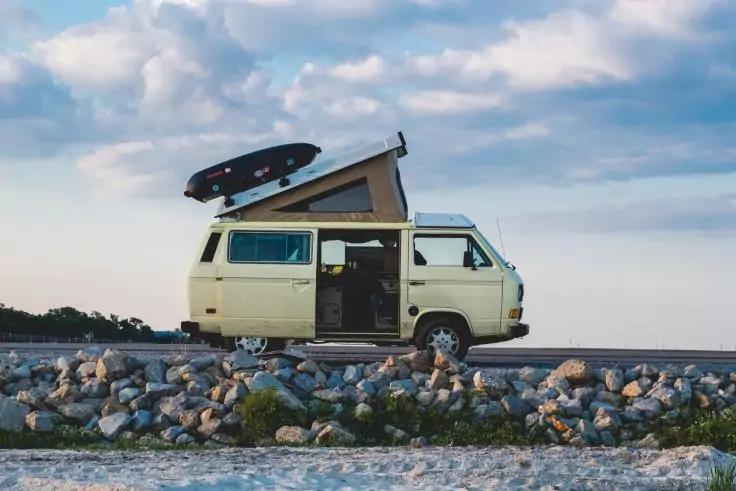 Hellbeiger Camper-Van mit aufgeklapptem Aufstelldach und Surfboard auf dem Dach, geparkt am felsigen Ufer, Seitent&uuml;r offen, Meer und wolkiger Himmel im Hintergrund