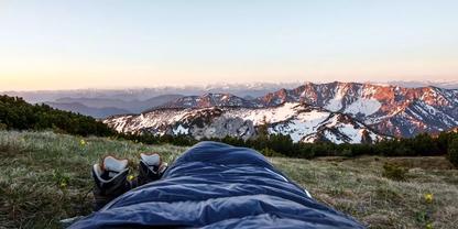 Blick aus einem Schlafsack auf eine grasbewachsene Wiese, Bergschuhe neben dem Schlafsack, im Hintergrund schneebedeckte Berge und ein Himmel mit oranger Horizontlinie