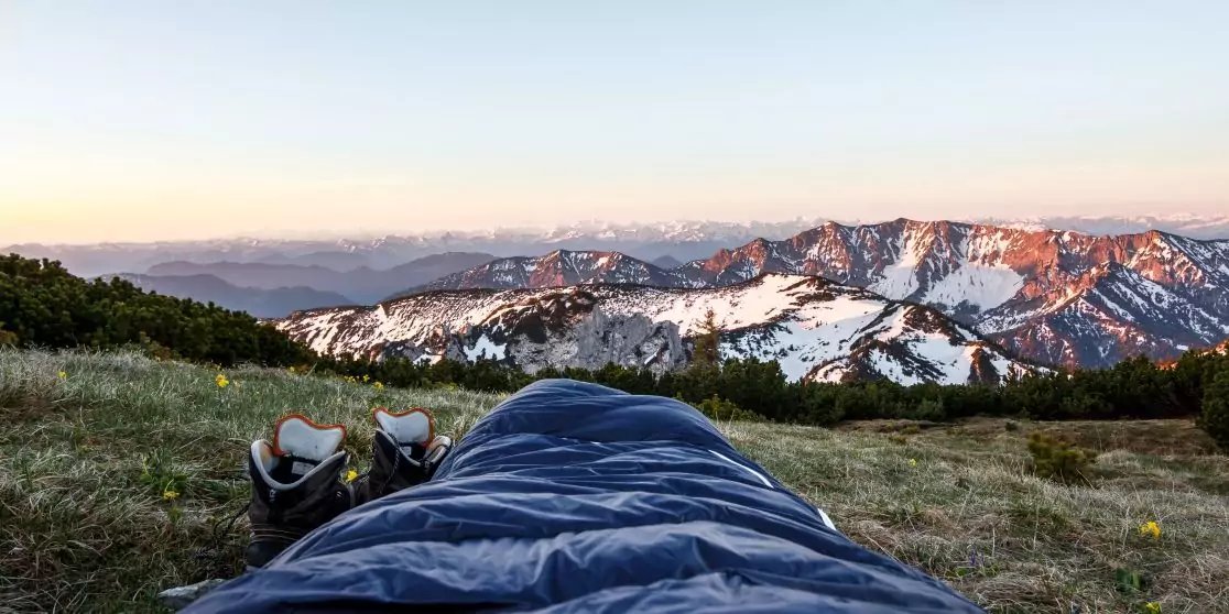 Blick aus einem Schlafsack auf eine grasbewachsene Wiese, Bergschuhe neben dem Schlafsack, im Hintergrund schneebedeckte Berge und ein Himmel mit oranger Horizontlinie
