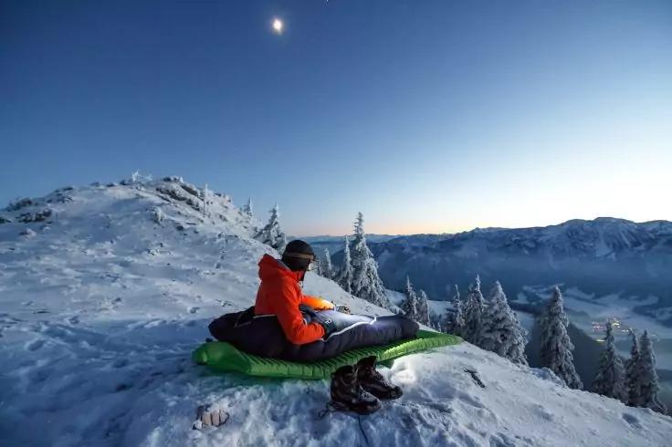 Person in oranger Jacke sitzt auf einer gr&uuml;nen Isomatte auf einem schneebedeckten Berggrat; Stiefel daneben; schneebedeckte B&auml;ume und entfernte Gipfel unter klarem Himmel mit Mond