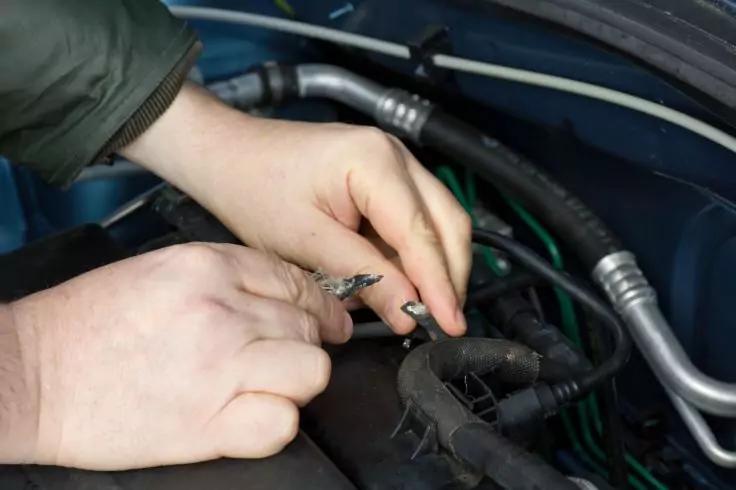 Zwei Hände halten freiliegende Drähte und Kabel neben Schläuchen und Bauteilen im Motorraum eines Autos