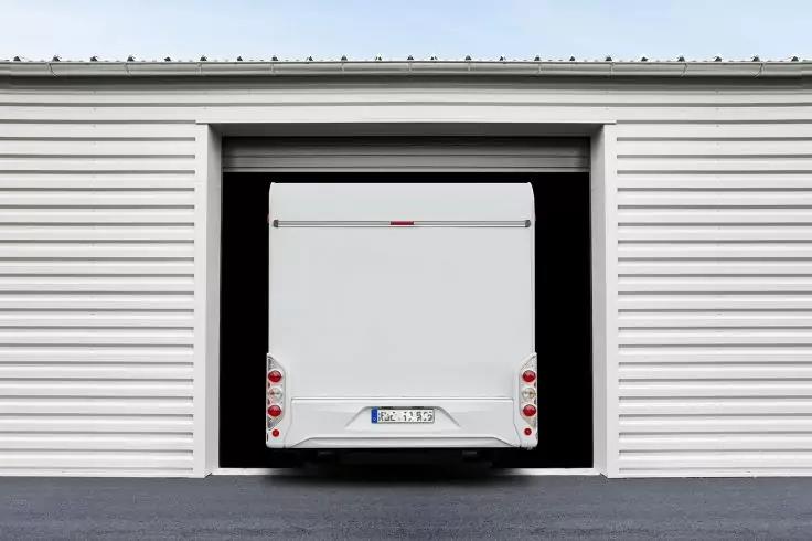 Rückansicht eines weißen Lieferwagens, der in einem geöffneten Lagergaragentor steht; seitliche weiße Metallwände mit horizontalen Profilen, dunkler Innenraum und Asphaltboden