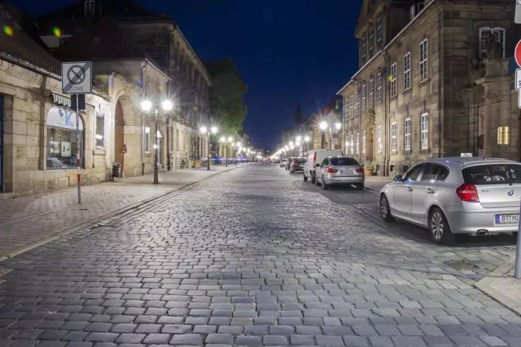 Kopfsteinpflasterstraße bei Nacht, parkende Autos auf der rechten Seite, beleuchtete Straßenlaternen und Gebäude beidseits