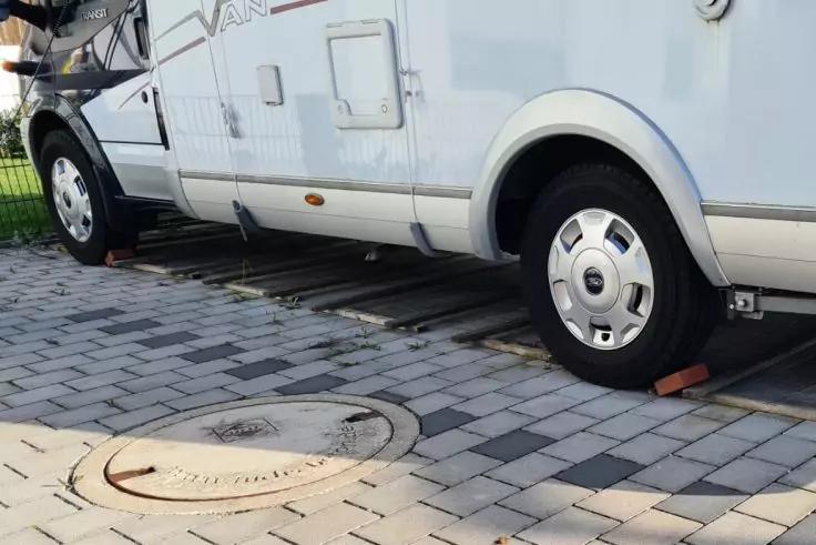 Seitenansicht eines weißen Wohnmobils auf grauem Pflaster; zwei Reifen stehen auf Holzkeilen; im Vordergrund ein runder Kanaldeckel; Zaun und Rasen im Hintergrund