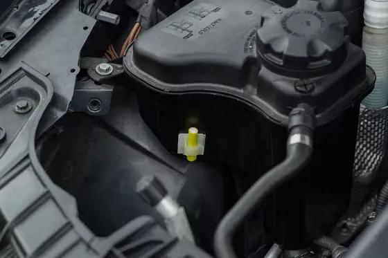 Motorraum, schwarzer Ausgleichsbehälter mit Schraubdeckel, umliegende Kunststoffverkleidung und kleiner gelber Schlauch-/Klemmclip