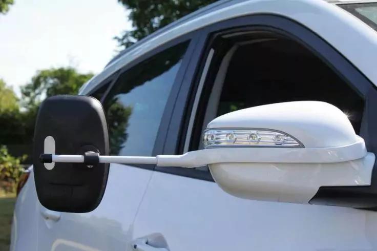 Wei&szlig;es Auto, sichtbare Beifahrert&uuml;r und Au&szlig;enspiegel; an diesem eine rechteckige schwarze Zusatzspiegelvorrichtung an einer silbernen Stange befestigt; B&auml;ume im Hintergrund