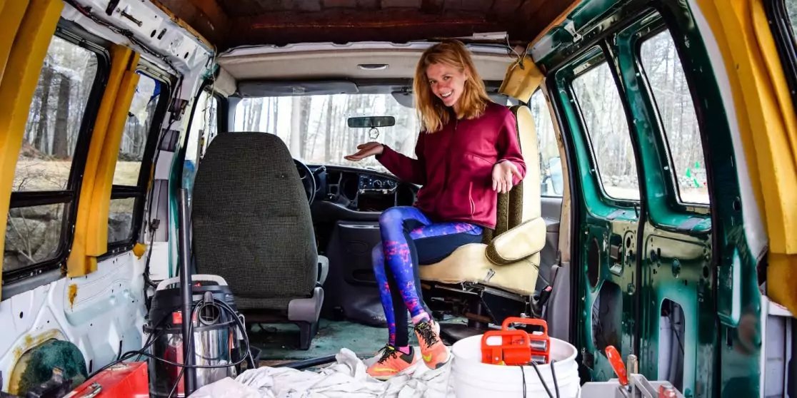 Person mit blondem Haar sitzt in einem offenen Lieferwagen, tr&auml;gt bordeauxroten Pullover und blaue gemusterte Leggings, barfu&szlig;; im Innenraum sind Sitze, freiliegende Innenverkleidung, Kabel, ein oranger Generator, ein roter Kanister, eine wei&szlig;e Decke