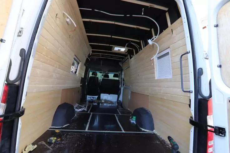 Innenraum eines geöffneten weißen Transporters mit hellen Holzplatten an den Seiten, sichtbaren Deckenbalken, schwarzem Boden mit Metallleisten, offenen Hecktüren, losen Kabeln und Werkzeugen