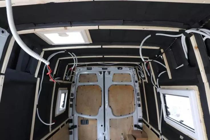Innenraum eines Fahrzeugs mit hinteren Metalldoppeltüren, Holzplatten an den Türen, schwarze Isolierplatten an Wänden und Decke, mehrere weiße Kabel entlang der Decke, rechteckiges Dachfenster, zwei seitliche Fenster, Holzfußboden