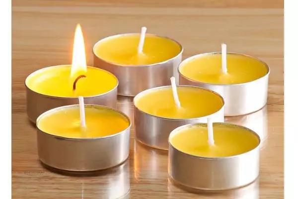 Sechs gelbe Teelichter in Metallhüllen auf einer Holzoberfläche, eines brennt