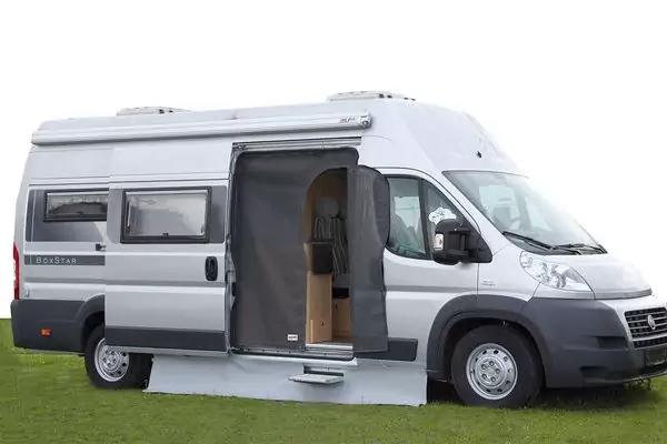 Weißes Wohnmobil mit geöffneter Seitentür, sichtbares Holzinterieur, ausgestellte Fußmatte auf Gras