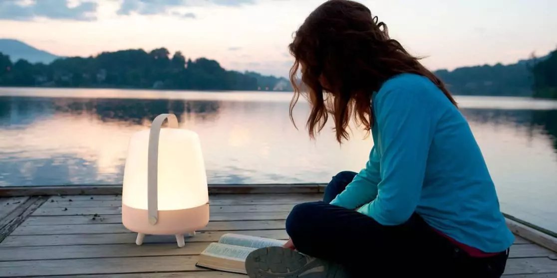 Person sitzt auf einem Holzsteg am See neben einer wei&szlig;en, leuchtenden Laterne mit Griff, im Hintergrund Wasser, Baumreihe und Himmel