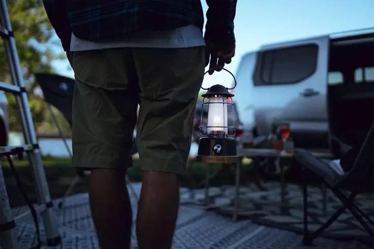 Unterk&ouml;rper einer Person in Shorts, die eine Laterne h&auml;lt; im Hintergrund Campingbus, Klappst&uuml;hle und ein kleiner Tisch