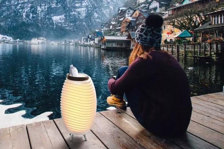 Person sitzt mit dem R&uuml;cken zur Kamera auf einem Holzsteg, tr&auml;gt eine Winterm&uuml;tze mit Bommel und einen dunklen Mantel; eine leuchtende Laterne steht auf dem Steg, ruhiger See, H&auml;user am Ufer und schneebedeckte Berge im Hintergrund