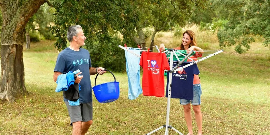 Ein Mann und eine Frau stehen auf einer Wiese unter B&auml;umen neben einem Drehw&auml;schetrockner mit aufgeh&auml;ngter Kleidung; der Mann h&auml;lt einen blauen W&auml;schekorb und Kleidung, die Frau h&auml;ngt ein Kleidungsst&uuml;ck auf