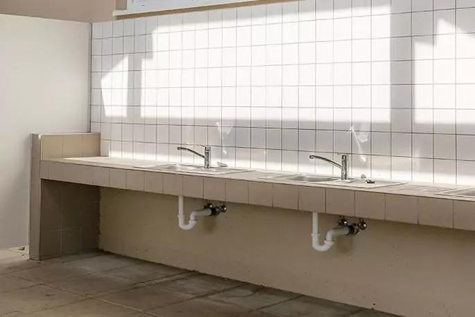 Zwei nebeneinander montierte Waschbecken mit Wasserh&auml;hnen und sichtbaren Abflussrohren an einer langen Arbeitsplatte vor einer wei&szlig;en, quadratisch gefliesten Wand