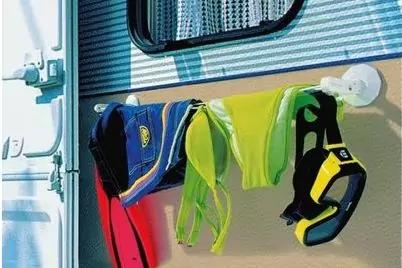 Drei Kleidungsst&uuml;cke h&auml;ngen an einer Wandhalterung neben einem Wohnwagenfenster: zwei neongelbe St&uuml;cke und ein dunkelblau-schwarzes Kleidungsst&uuml;ck mit gelben Akzenten