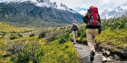 Zwei Wanderer mit Rucks&auml;cken gehen auf einem schmalen Bergpfad durch gr&uuml;ne Vegetation Richtung schneebedeckter Berge unter leicht bew&ouml;lktem Himmel