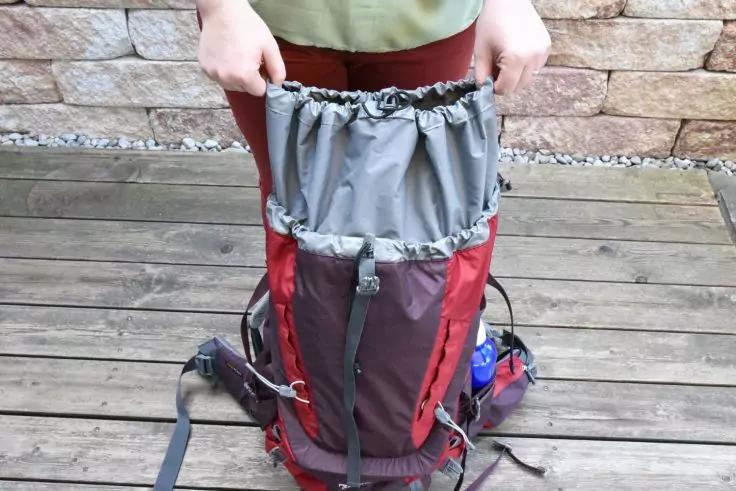 Roter und grauer Rucksack mit offenem Oberteil, von einer Person an den Seiten gehalten; steht auf Holzbohlen vor einer Steinmauer