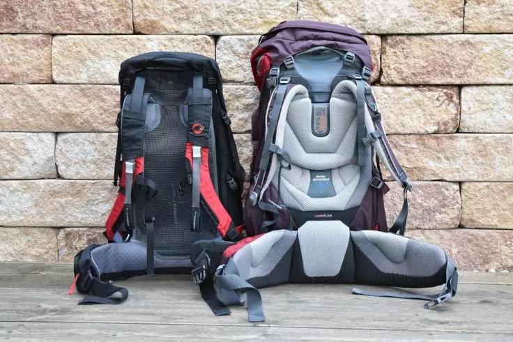 Zwei gro&szlig;e Rucks&auml;cke, einer schwarz mit roten Tr&auml;gern und einer lila-grau; R&uuml;cken- und H&uuml;ftgurte sichtbar, auf Holzboden vor einer Steinwand