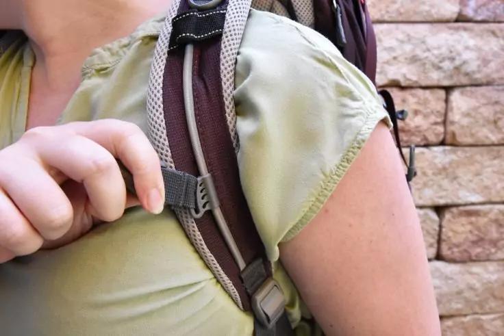 Nahaufnahme der Schulter und Brust einer Person, eine Hand h&auml;lt den Rucksackgurt; olivgr&uuml;nes T-Shirt und Steinmauer im Hintergrund