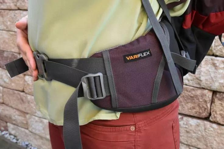 Nahaufnahme eines Oberk&ouml;rpers mit olivgr&uuml;nem Oberteil und roten Hosen; gepolsterter H&uuml;ftgurt eines Rucksacks mit Aufschrift 'VARIFLEX', Hand h&auml;lt den Gurt, steinerne Mauer im Hintergrund