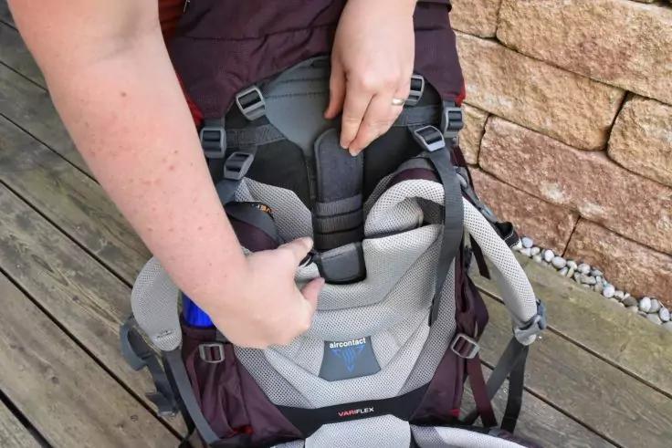 H&auml;nde einer Person justieren einen Gurt an der grauen, gepolsterten R&uuml;ckenpartie eines dunkelroten Rucksacks auf einem Holzdeck vor einer Steinmauer; Ring am rechten Ringfinger sichtbar