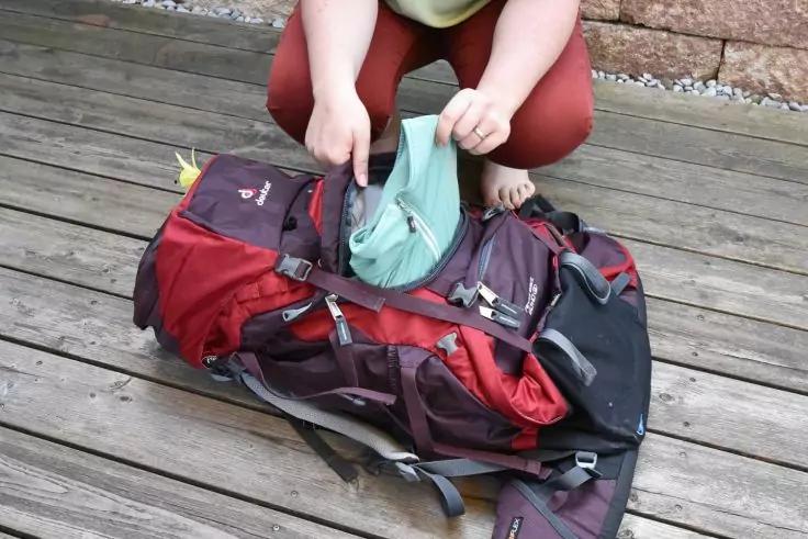 Person kniet auf Holzdeck und steckt ein hellgr&uuml;nes Kleidungsst&uuml;ck in einen ge&ouml;ffneten roten und schwarzen Rucksack