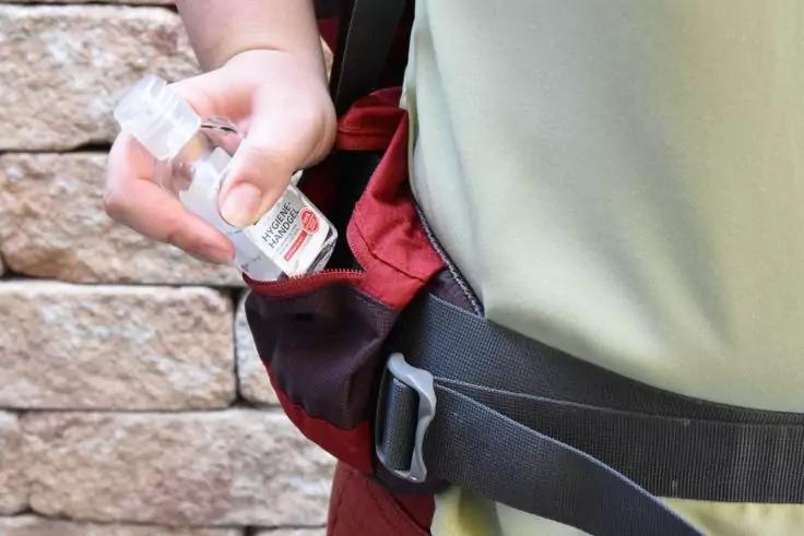 Hand einer Person steckt eine kleine transparente Pumpflasche mit Etikett in eine rote H&uuml;fttasche; Person tr&auml;gt gr&uuml;nes Shirt und schwarzen Gurt; Steinmauer im Hintergrund