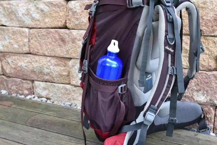 Rucksack mit grauen Tr&auml;gern vor einer Sandsteinmauer; blaue Trinkflasche in der Seitentasche