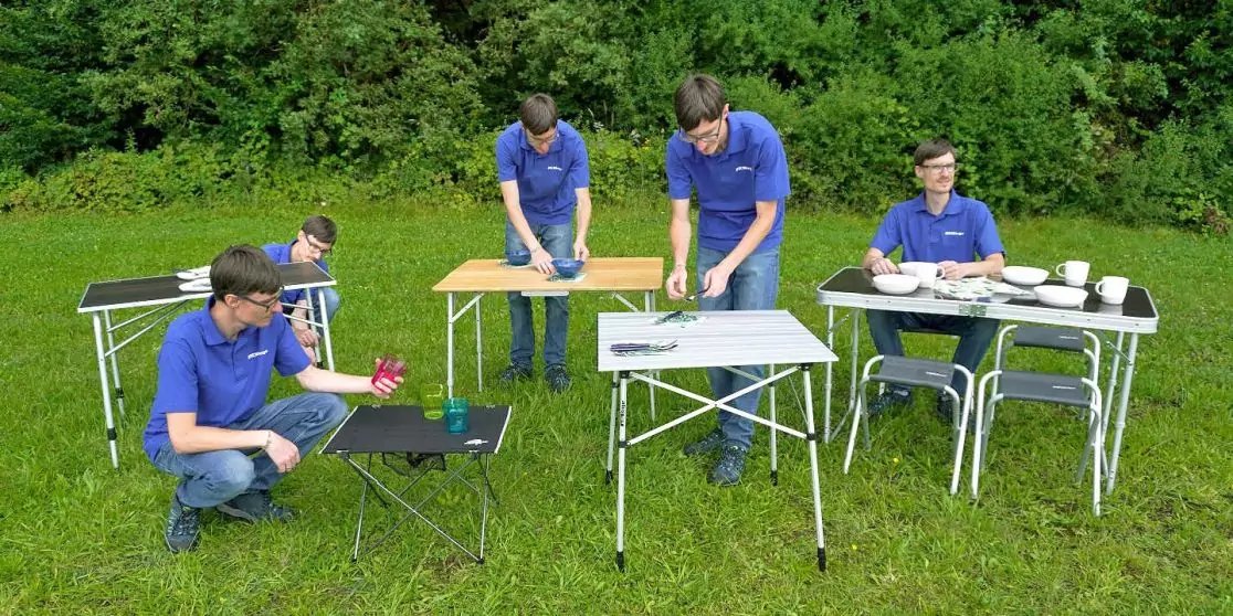 F&uuml;nf Personen in blauen Poloshirts an mehreren Klapptischen auf einer Wiese vor B&auml;umen; auf den Tischen eine Topfpflanze, Geschirr und kleine Gegenst&auml;nde