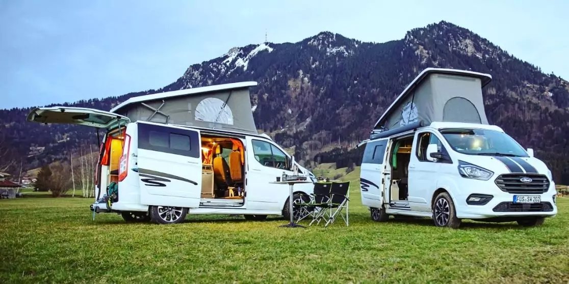 Zwei wei&szlig;e Campingbusse mit aufgeklappten Aufstelld&auml;chern auf einer Wiese, dazwischen Klapptisch und St&uuml;hle, im Hintergrund Berge