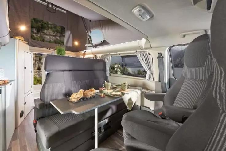 Innenraum eines Wohnmobils mit Tisch, Brot und Geschirr auf dem Tisch, einer Sitzbank, einem drehbaren Fahrersitz, Gardinen und Schr&auml;nken
