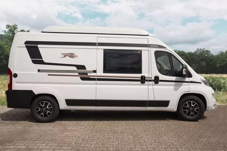 Wei&szlig;es Fahrzeug mit Hochdach und seitlichem Fenster, geparkt auf gepflasterter Fl&auml;che; schwarze Dekorstreifen, zwei R&auml;der sichtbar; im Hintergrund Gras und bew&ouml;lkter Himmel