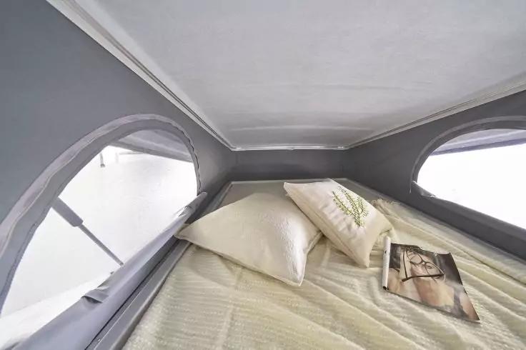 Innenraum mit Matratze, zwei Kissen (eins mit Blattmuster), einer Zeitschrift und drei Netzfenstern, graue Stoffw&auml;nde und -decke