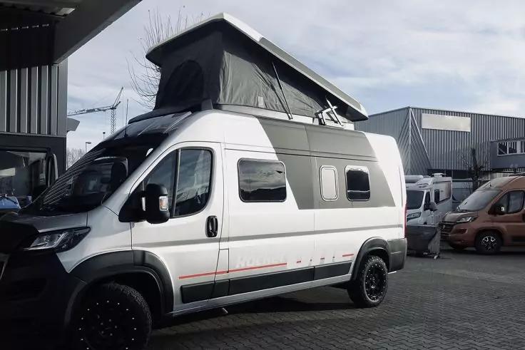 Wei&szlig;er Kastenwagen mit aufgeklapptem schwarzem Aufstelldach, drei Seitenfenster, schwarze Felgen, geparkt auf gepflastertem Hof vor Industriegeb&auml;uden; weitere &auml;hnliche Fahrzeuge im Hintergrund, bew&ouml;lkter Himmel