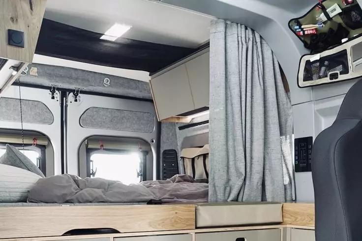 Innenraum eines Campers mit Holz-Liegeplattform, grauer Bettw&auml;sche und Kissen, Schubladen unter der Liege, graue Vorh&auml;nge, zwei Heckt&uuml;ren mit Fenstern, Deckenleuchte und H&auml;ngeschrank