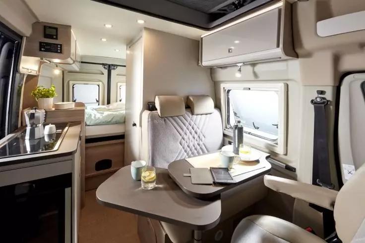 Innenraum eines Wohnmobils mit Esstisch, zwei gepolsterten Sitzen, Weinflasche, zwei Gl&auml;sern, Tellern, K&uuml;chenzeile mit Sp&uuml;le, Oberschr&auml;nken und Bett im Heck