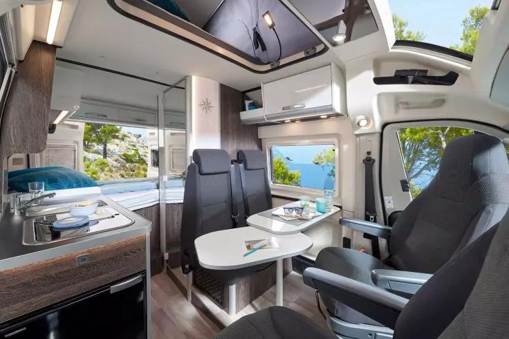 Innenraum eines Wohnmobils mit zwei drehbaren Vordersitzen, Tisch mit Geschirr, Sitzbank mit blauen Kissen, K&uuml;chenzeile mit Sp&uuml;le und Herd, Oberschr&auml;nken, Fenster mit Blick auf Meer und B&auml;ume, Dachfenster